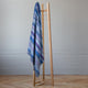 Morona baby alpaca wool blanket – a wooden coat rack stands upright, holding a multicolored, striped fabric draped over it. The scene is set against a plain, light-colored wall with a patterned wood floor