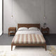 Quijos alpaca wool blanket – a wooden bed with a striped blanket and pillows is centered between two wooden nightstands under pendant lights. The setting is a minimalist bedroom with gray walls and flooring