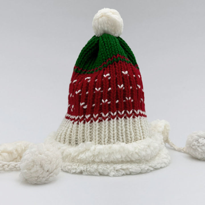 A knitted beanie displays a red and green pattern with a white pom-pom on top, sitting on a flat surface with a fuzzy white brim and long tassels.