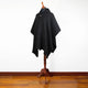 A black poncho with a hood and front pocket is displayed on a wooden mannequin stand, set against a simple white wall and wood floor background.
