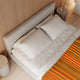 Putumayo baby alpaca wool blanket – a neatly made bed with white pillows and beige sheets features a striped orange and brown blanket. Situated against a wooden paneled wall, a round bedside table holds a glass vase