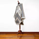 A gray and beige patterned poncho with fringe is displayed on a wooden stand, set against a hardwood floor and plain white background.