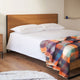 Anzu baby alpaca wool blanket – a neatly made bed with white pillows and a colorful plaid blanket is in a minimalist bedroom with wooden flooring and a small shelf holding a blue vase