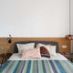 Imbabura baby alpaca wool blanket – a bed with a gray headboard and striped bedding, adorned with patterned pillows. A wooden side table with dried flowers and books sits beside it, under modern wall lights