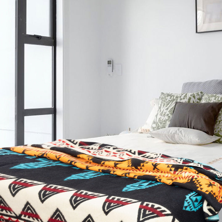 A bed is covered with a colorful quilt featuring geometric patterns. It is adorned with multiple cushions in a bright, white-walled room beside a tall window and artwork.