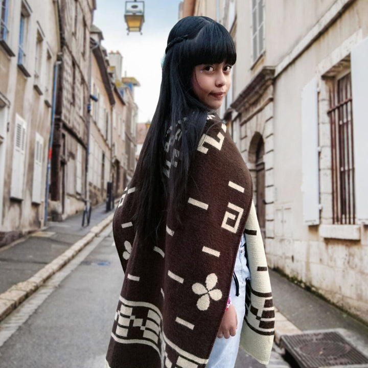 Kids Clint Eastwood alpaca wool poncho – a person wrapped in a patterned poncho stands on a narrow, cobblestone street lined with old buildings. The person looks over their shoulder in an urban, historical setting