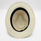 A beige woven hat lies upside-down, revealing a black inner band and a textured inner crown. It rests against a plain, light background.