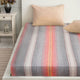 Ajilanga baby alpaca wool blanket – a bed is covered with a striped sheet featuring grey, pink, and orange hues. Two beige pillows are placed at its head. Nearby, there's a white nightstand with a vase and lantern