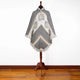 A poncho with geometric patterns hangs on a wooden mannequin stand. The poncho features fringed edges and earth-toned designs. This setup is placed on a wooden floor against a plain white background.