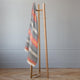 Ajilanga baby alpaca wool blanket – a wooden blanket rack holds a striped gray, orange, and white blanket, positioned on a wood floor against a light gray wall