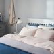Bed with pastel sheets and multiple pillows is neatly made; nearby, a white lamp and simple decor on a gray dresser complete the bright, minimalist bedroom setting.