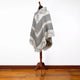 A patterned poncho drapes over a wooden mannequin stand, featuring geometric designs in gray and white tones. It rests on a wooden floor against a plain white background.