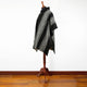 A black and white patterned poncho drapes over a wooden mannequin in a well-lit room with wooden flooring, set against a plain white wall.