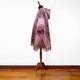 A hooded poncho with geometric patterns and fringed edges is displayed on a wooden mannequin stand, set against a plain white wall and wooden floor.