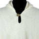 A cream-colored knit sweater features a single wooden button fastening near the collar. The garment is displayed on a black mannequin backdrop.