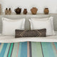 Sucua baby alpaca wool blanket – two white pillows rest on a bed with a striped blanket and patterned bolster cushion. Ancient-style pottery decorates a minimalist shelf above the bed