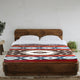 Qhusi extra large alpaca wool blanket – a wooden bed features a colorful geometric-patterned blanket. It's placed in a minimalistic room with white walls, a small potted plant, and a round wooden side table