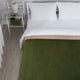 A neatly made bed features white pillows and sheets, covered by a beige and green blanket. A white nightstand beside it holds books and a lamp in a minimalist room.