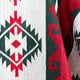 Intricate textile displaying geometric patterns, predominantly red, green, and white, with symmetrical diamond shapes. The fabric folds slightly, highlighting its woven texture.