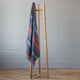 Huasaga baby alpaca wool blanket – a wooden clothing rack holds a colorful, striped blanket draped over it. It stands on a patterned wooden floor against a light gray wall, creating a minimalist interior setting