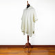 A cream-colored poncho rests on a wooden mannequin stand, displayed in a room with a polished wooden floor and a plain white wall background.