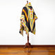 A colorful poncho with geometric patterns drapes on a wooden mannequin stand, set against a plain white wall and polished wooden floor.