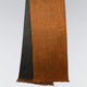 A two-toned scarf hangs vertically, featuring a burnt orange and dark gray color scheme with fringed ends, set against a plain light gray background.