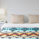 A bed with beige pillows is adorned with a colorful blanket featuring turquoise geometric patterns. It sits in a minimalist bedroom with a white lamp and nightstand.