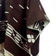 Kids Clint Eastwood alpaca wool poncho – brown and white poncho drapes gracefully, featuring geometric patterns and symbols. The design includes lines and a floral motif, with fringes at the bottom, set against a plain background