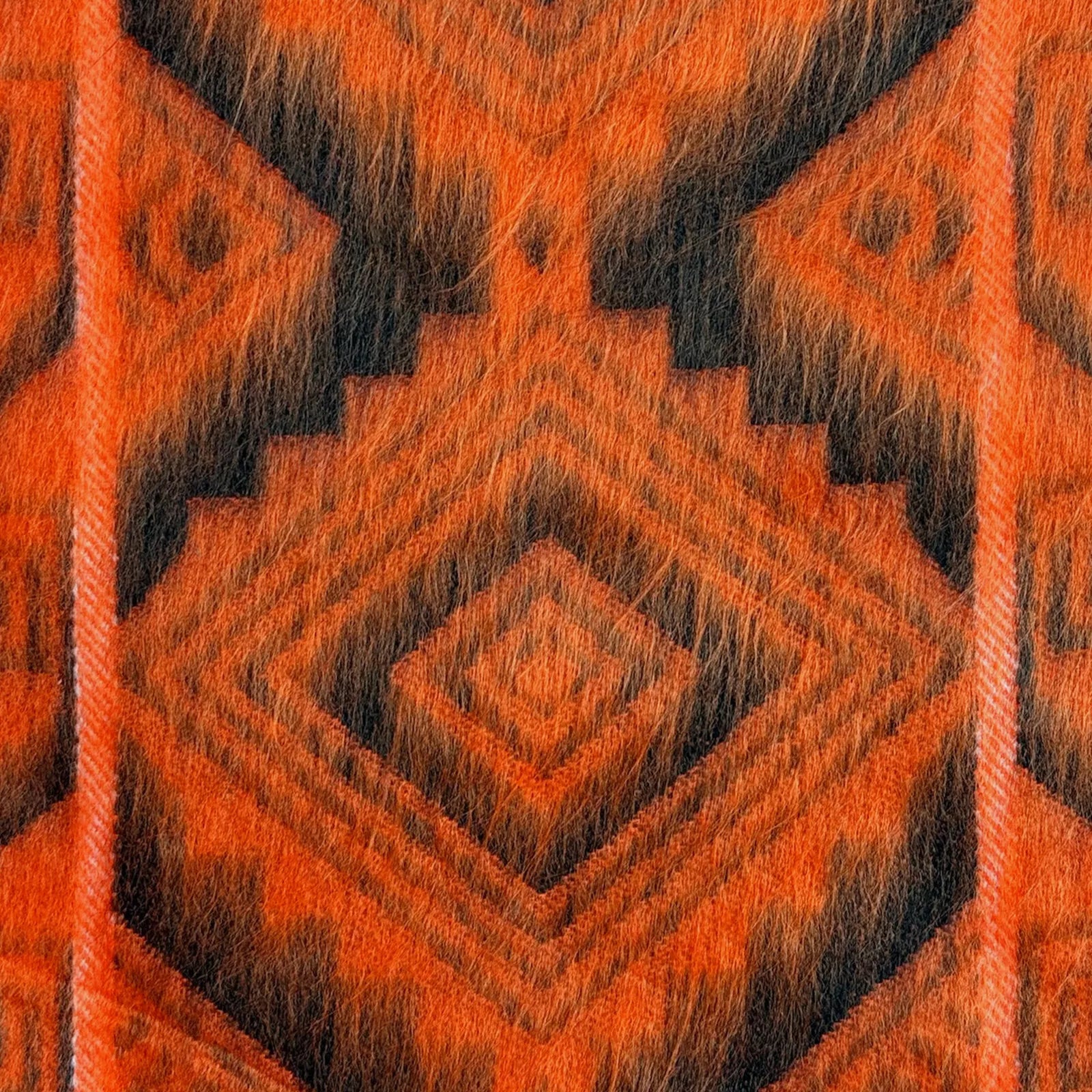 A close-up of an orange and black woven fabric displaying a geometric pattern with overlapping diamond shapes, creating a striking, symmetrical design. The texture appears soft and fuzzy.