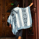 A person in a patterned poncho, wearing a black hat, pushes open a wooden door. They stand in an ornately decorated entrance with a visible ENTRADA sign in the background.