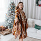 A person wearing a patterned poncho stands beside a decorated Christmas tree in a cozy living room with a white sofa. A green pillow reads Joyful.