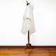 A white poncho with a hood is displayed on a wooden mannequin stand, set in a room with a wooden floor and a plain white wall background.