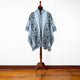 A wool poncho with intricate geometric patterns drapes over a wooden mannequin. The setting is a room with polished wooden floors and a plain white wall.