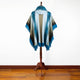 A blue and gray shawl with fringed edges is displayed on a wooden stand, set against a wooden floor and a plain white background.
