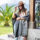 Zuleta llama wool hooded poncho – a person wearing a hat and gray poncho leans against a wooden post, set amidst greenery and a palm tree in the background