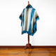 A poncho displayed on a wooden mannequin stand features blue and beige patterns with fringe edges, against a plain white background on a wooden floor.