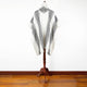 A white wool poncho with black, diamond-patterned stripes hangs on a wooden stand. The setting is a simple room with a wooden floor and a plain white wall.