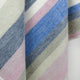 Yunga baby alpaca wool blanket – a woolen fabric features diagonal stripes in muted pink, blue, cream, and gray, creating a soft, pastel appearance. The texture is woven, providing a cozy, warm feel