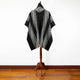 A black and white patterned poncho drapes over a wooden stand in a room with wooden flooring against a white wall.