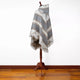A patterned gray and beige poncho with fringe edges is displayed on a wooden stand in a room with a hardwood floor and white walls.