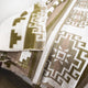 A woven blanket features intricate geometric patterns in olive green and white, draped over a surface, highlighting its detailed craftsmanship against a neutral background.