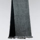A two-toned scarf, with black on one side and gray on the other, hangs vertically. The fringed ends add texture, set against a simple gray background.