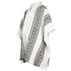 A white poncho with black diamond patterns hangs neatly, displaying fringe at the hem. It features a short collar with two decorative buttons, situated against a plain white background.
