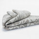A neatly folded, patterned blanket lies on a plain white surface, featuring a subtle, repetitive design in beige and light gray hues.