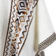 A white woven fabric showcases zig-zag and diamond patterns in brown and black on its border, draped elegantly in a softly lit, neutral setting.