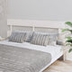 A neatly made bed with striped throw pillows rests against a light-colored headboard. A patterned blanket covers the bed. A potted plant decorates the minimalist, light-colored room.