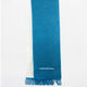 A blue woven scarf hangs vertically with fringed ends and a small geometric pattern near the bottom. It contrasts against a plain white background.