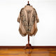A woven poncho with geometric patterns and fringe edges drapes over a mannequin, standing on a wooden floor against a white wall.