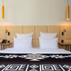 A neatly arranged bed features four white pillows and a geometric-patterned blanket. The bed is flanked by two wooden side tables, pendant lights, and reading lamps against a beige headboard.